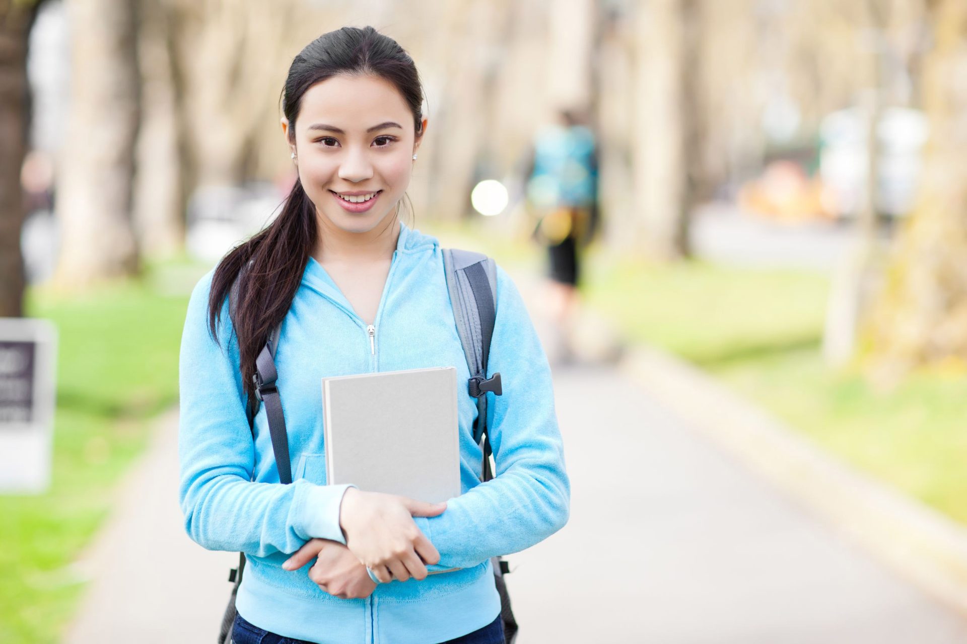 an Asian college student with a backpack and notebook smiles on a college campus