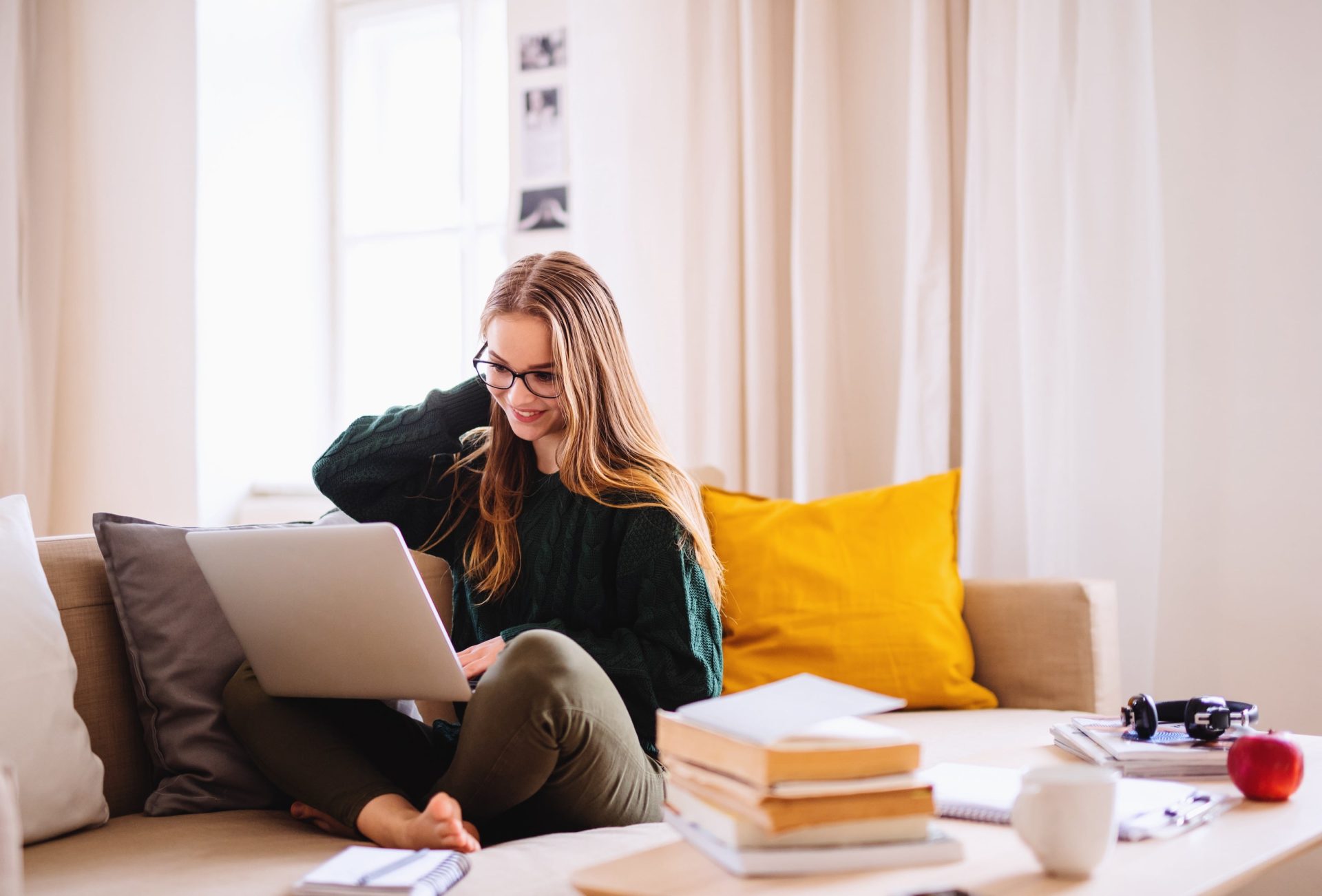a woman sits on a couch with her laptop and books smiling